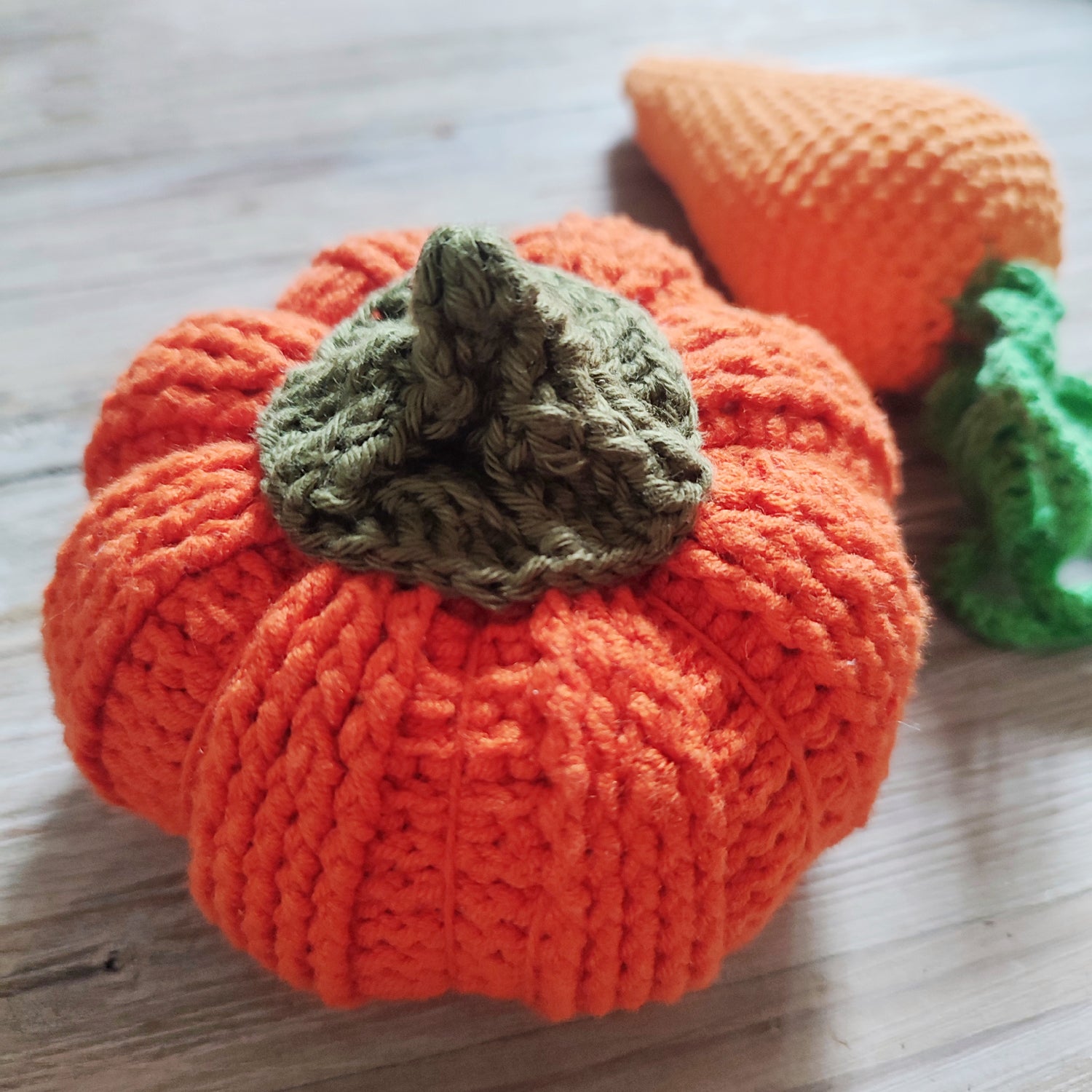 Citrouille en crochet orange avec une tige verte à côté d'une carotte en crochet sur une surface en bois.